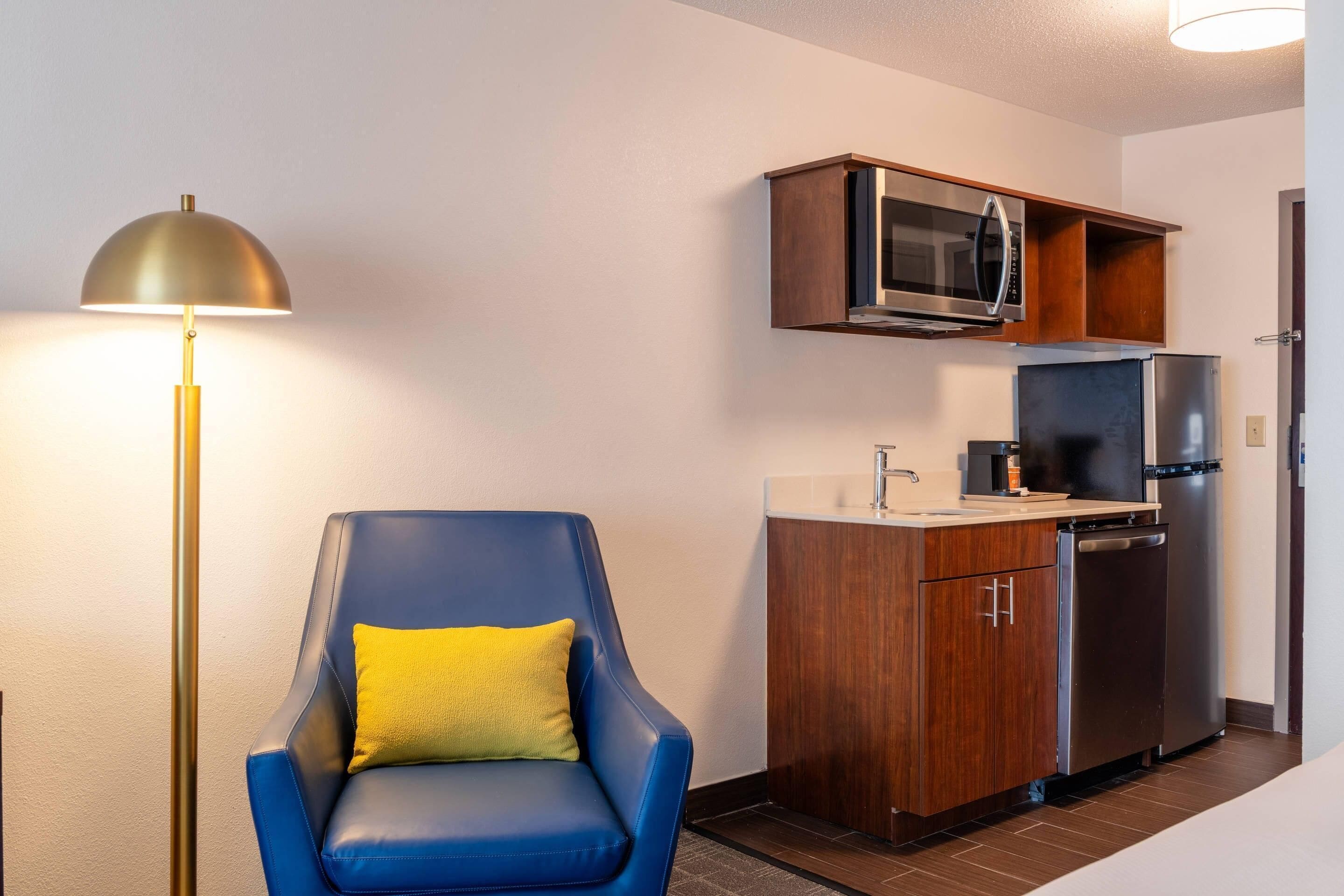 Una cocina compacta equipada con microondas, refrigerador y lavavajillas. Un sillón azul con un cojín amarillo se encuentra cerca de una lámpara de pie.