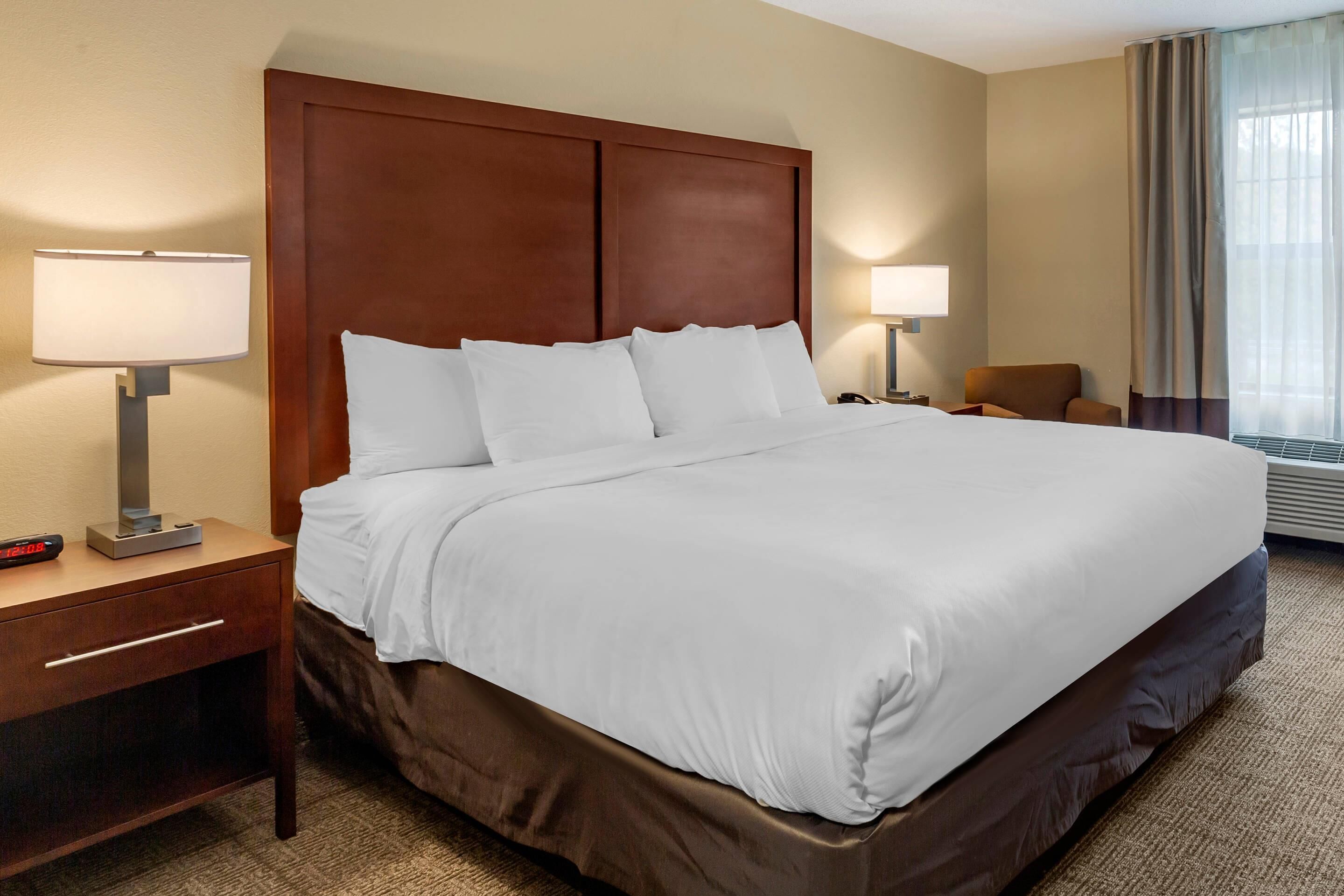 A neatly made hotel room with a large bed, two bedside tables with lamps, a chair, and a window with curtains.