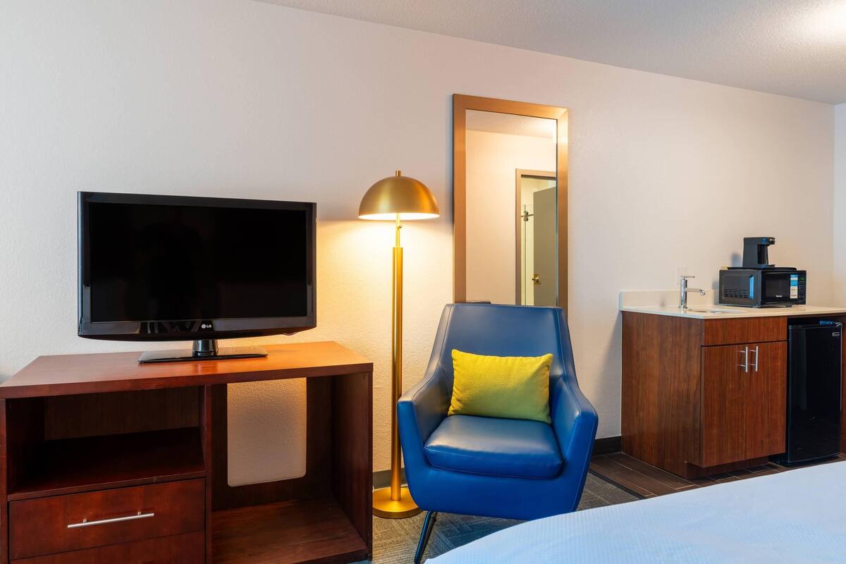 A hotel room with a TV, a desk, a chair, a lamp, a mirror, and a kitchenette.