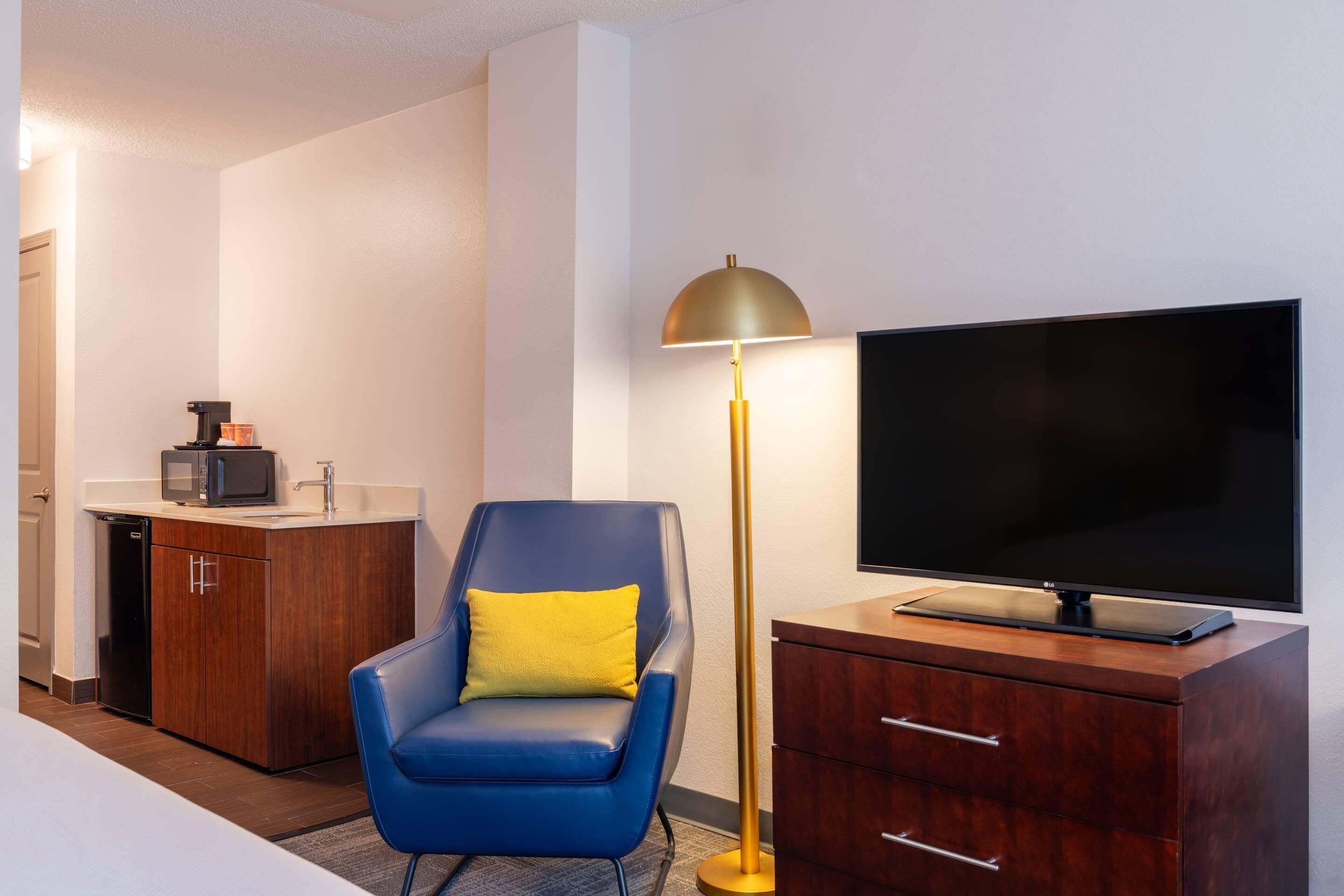 Een hotelkamer met een flatscreen televisie, een blauwe stoel met een geel kussen, een houten kast en een vloerlamp.