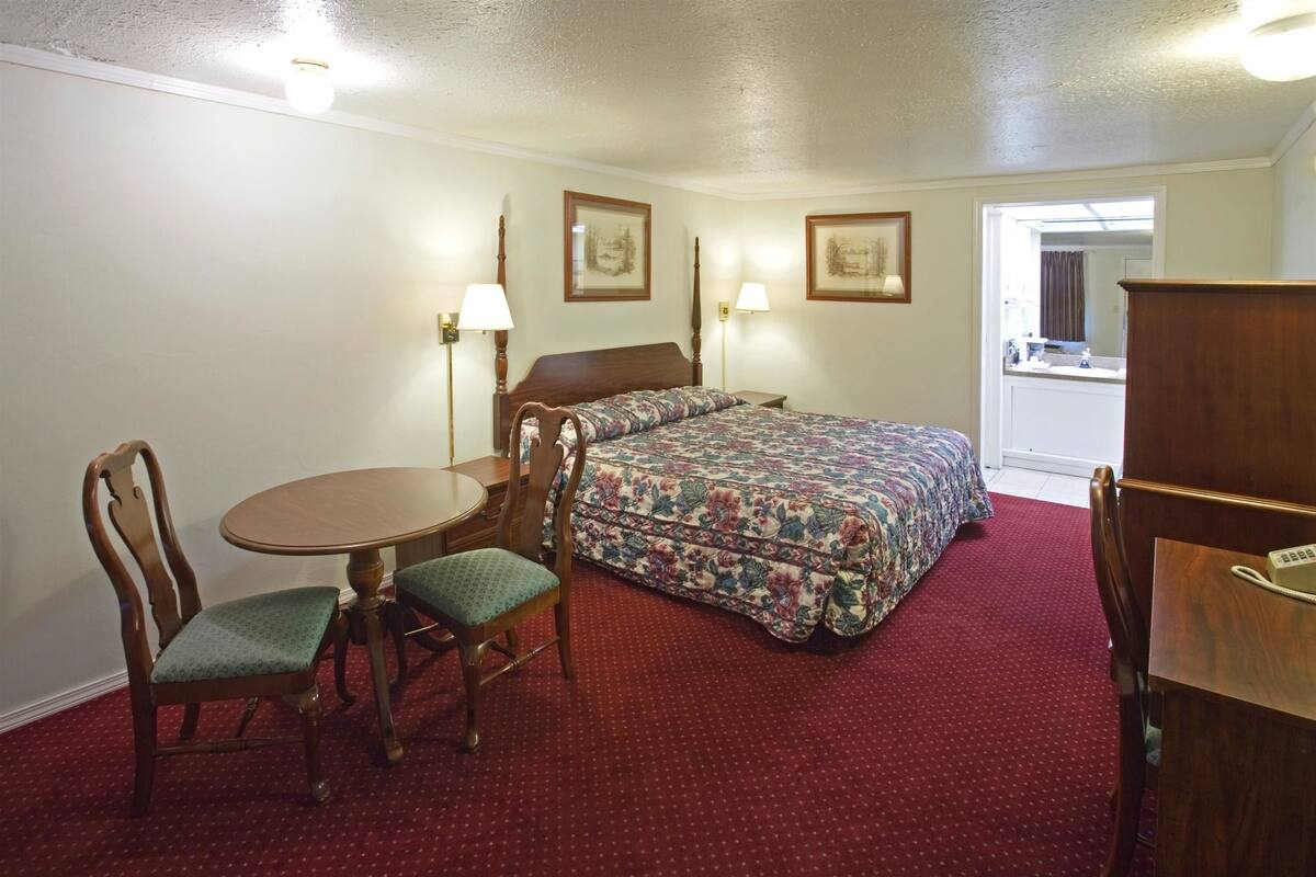 A hotel room with a bed, a desk, a chair, a table, and two lamps.