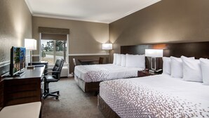 A hotel room with two beds, a desk, a chair, and a window with a view of trees.