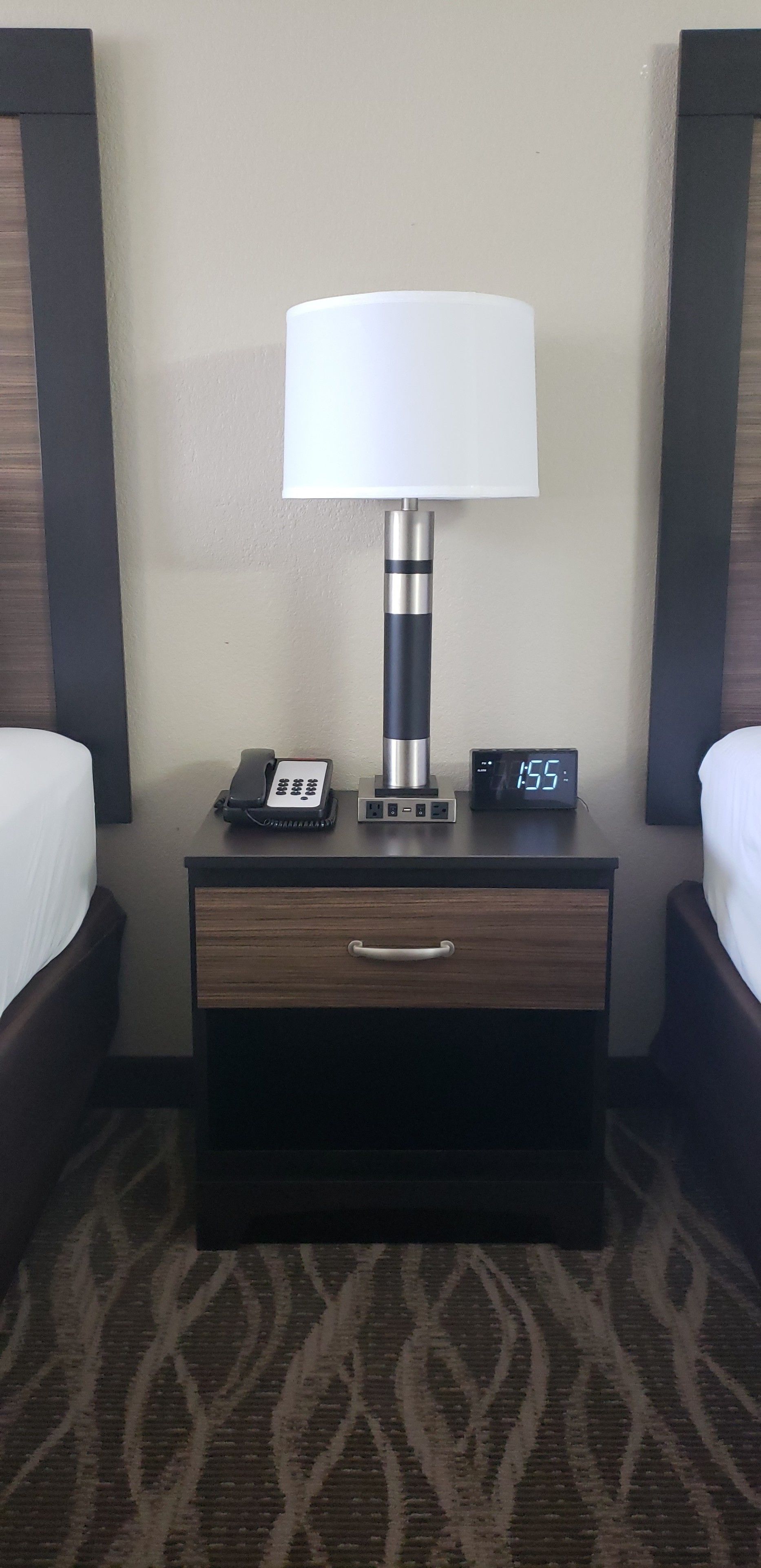 A hotel nightstand with a lamp, a digital clock displaying 15:55, a telephone, and a drawer.
