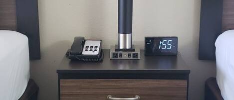 A hotel nightstand with a lamp, a digital clock displaying 15:55, a telephone, and a drawer.