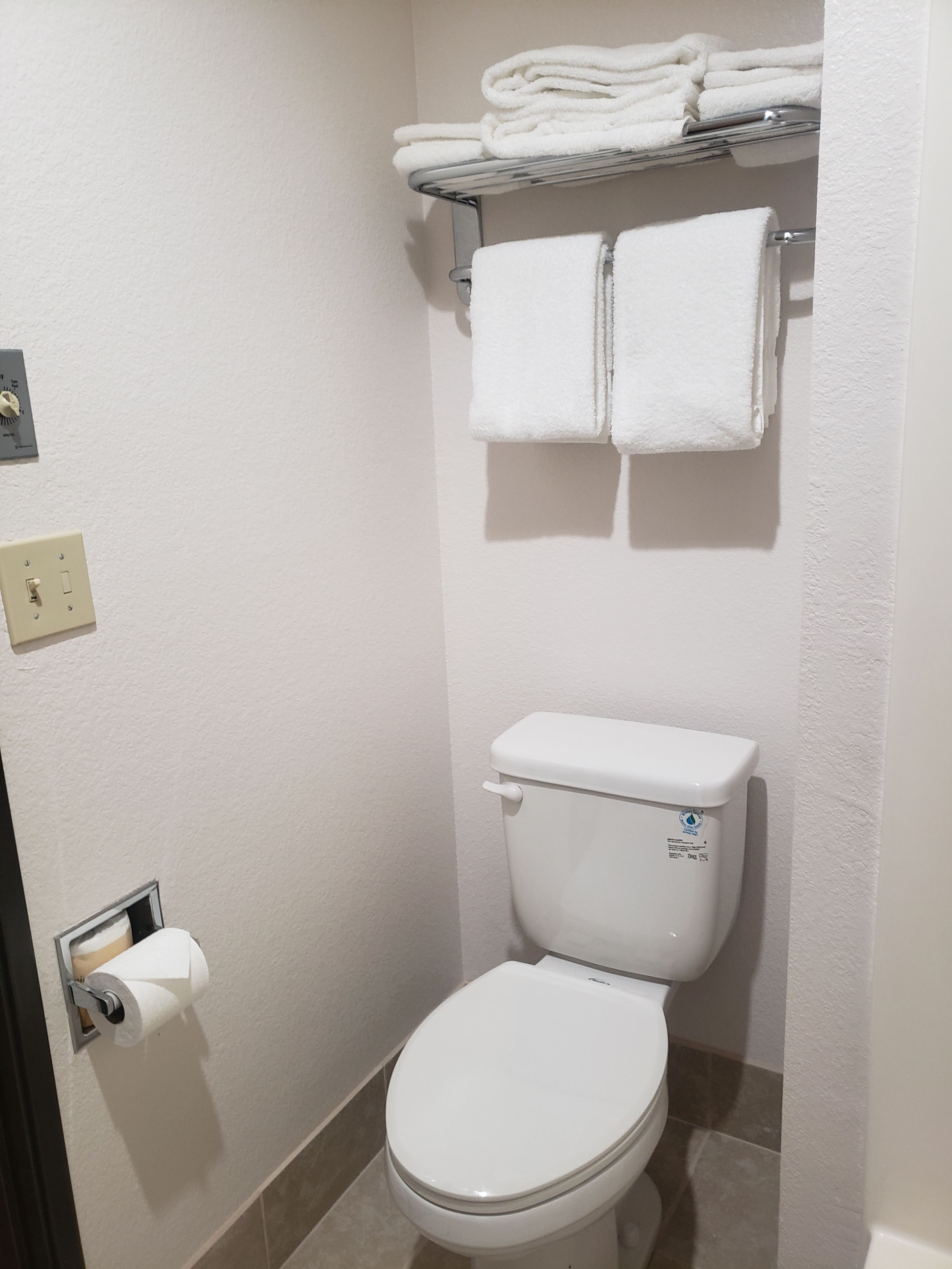 Un baño con inodoro, un perchero con toallas dobladas, dispensador de papel y un interruptor de luz en la pared.