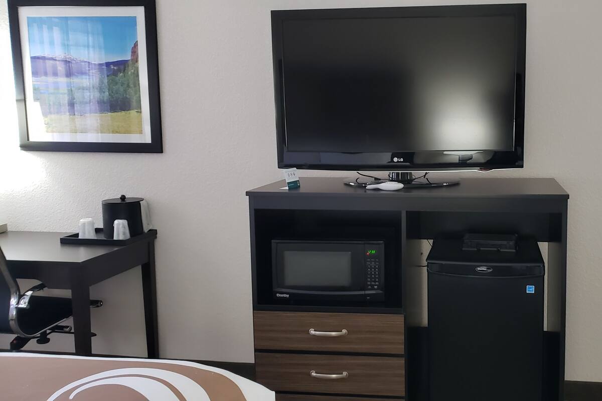 A hotel room with a flat-screen TV mounted on a black desk, a microwave, a small refrigerator, and a chair.