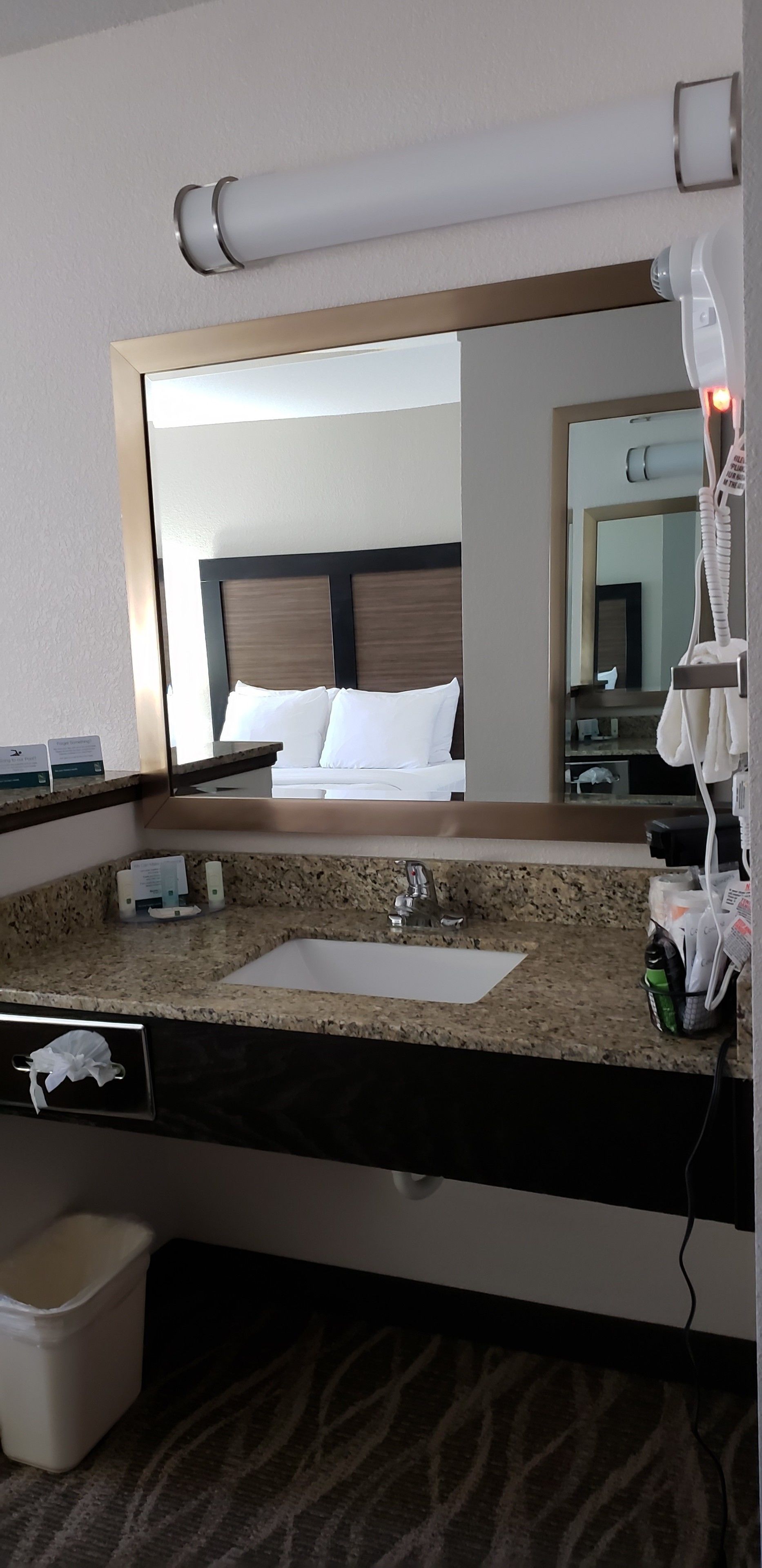 A hotel bathroom with a granite countertop, a sink, a mirror, a hairdryer, and a trash bin.