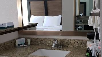 A hotel bathroom with a granite countertop, a sink, a mirror, a hairdryer, and a trash bin.
