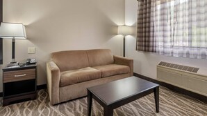 Habitación de hotel con sofá color beige, una mesita, una mesita de noche con lámpara y una ventana con cortinas de cuadros.