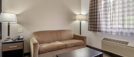 Ein Hotelzimmer mit einem beigen Sofa, einem kleinen Tisch, einem Nachttisch mit Lampe und einem Fenster mit Karottenvorhängen.
