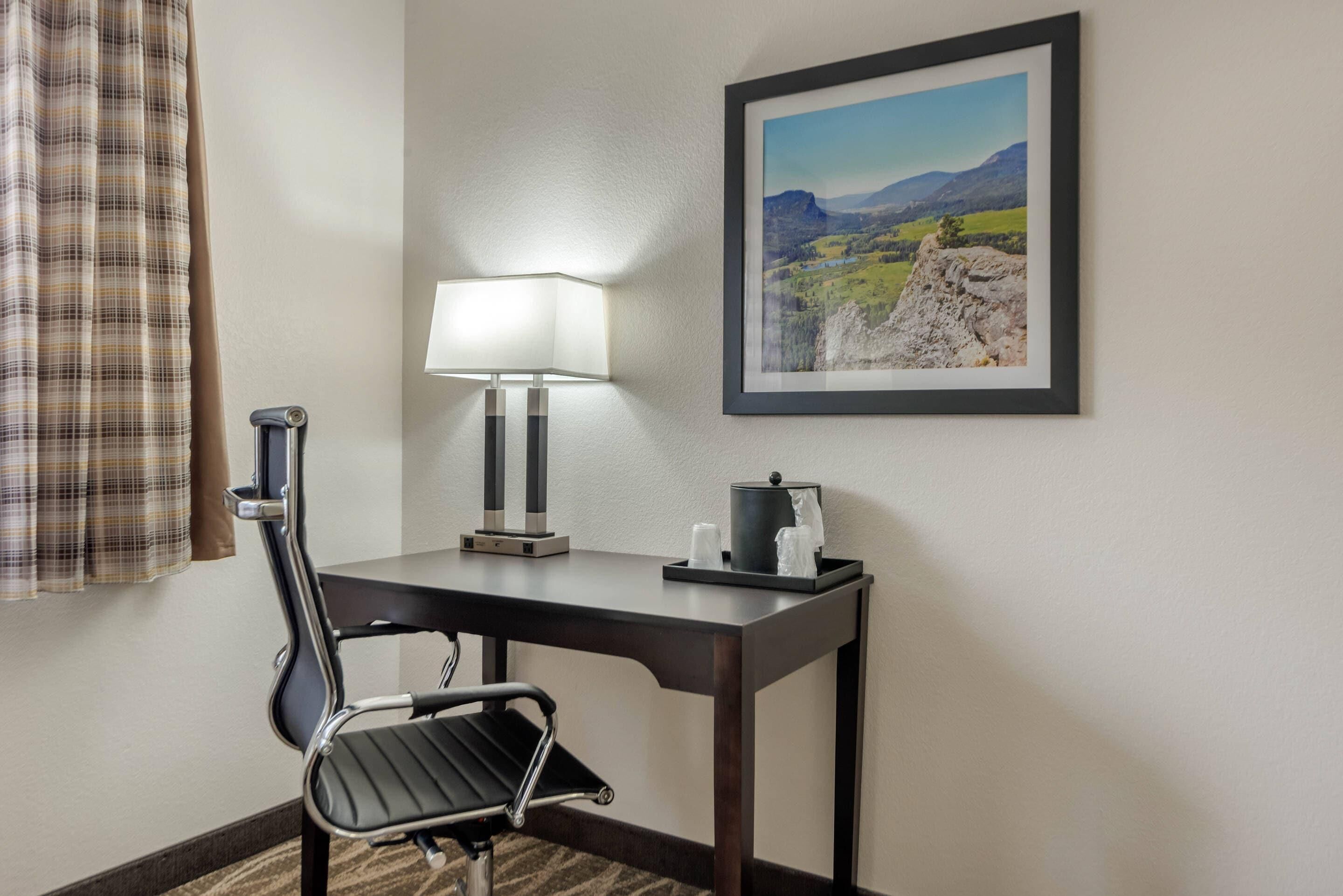 Een hotelkamer met een bureau, een stoel, een lamp, een lijst met een landschapsafbeelding, en een raam met gordijnen.
