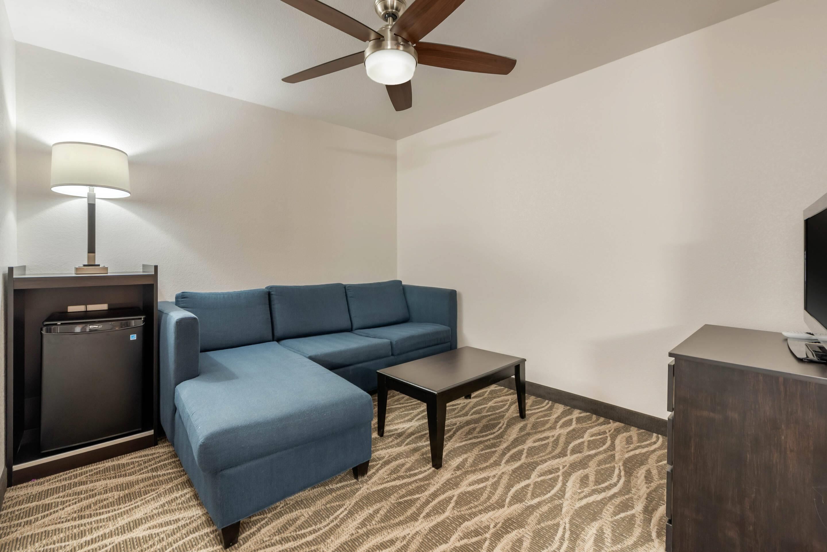 A hotel room with a blue sofa, a small table, a ceiling fan, a lamp, a TV, and a minibar.