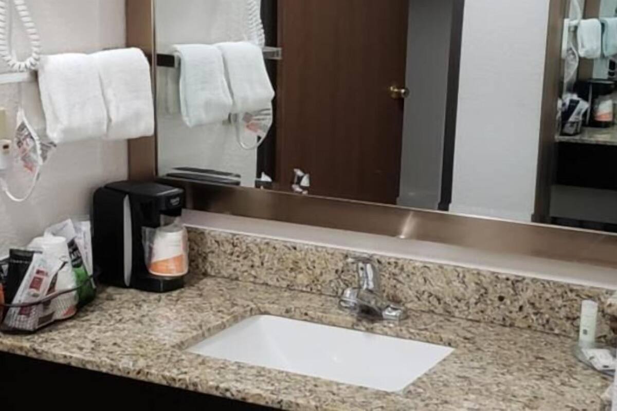 A hotel bathroom with a granite countertop sink, a coffee maker, and a mirror reflecting a door and towels.