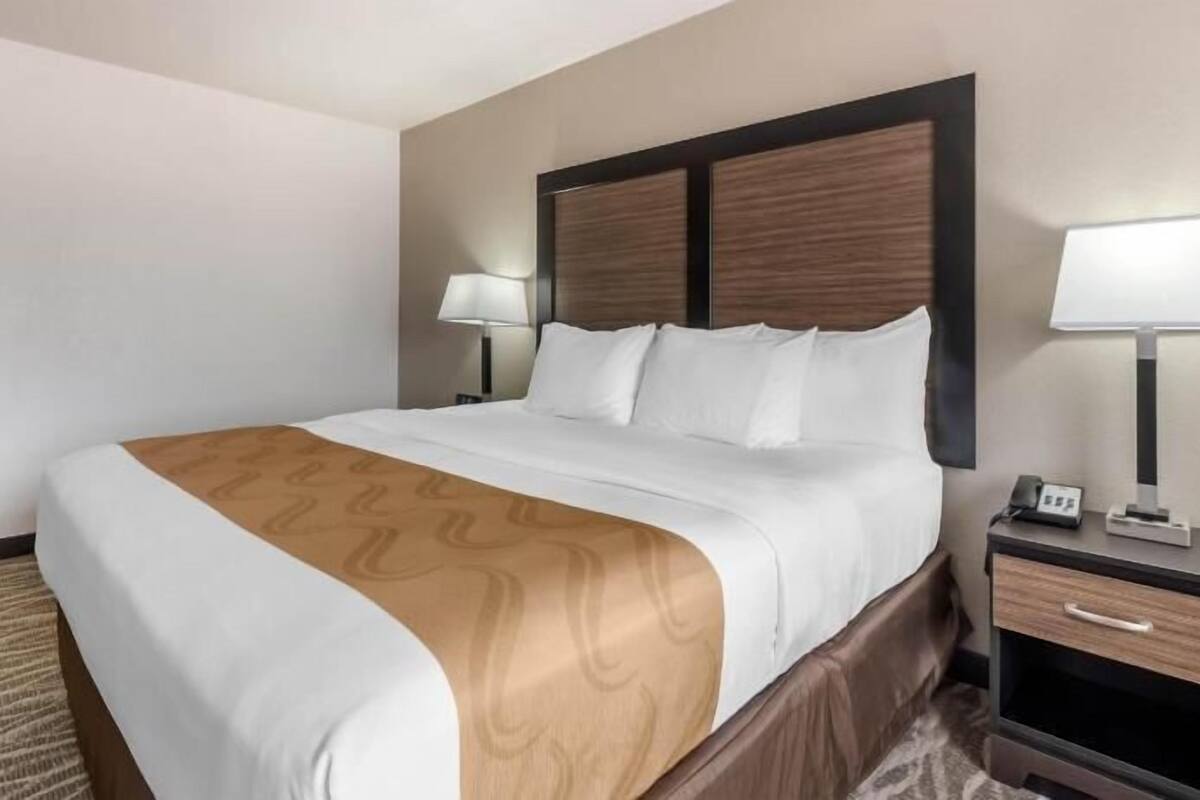 A hotel room with a large bed, a wooden headboard, two bedside tables with lamps, and a carpeted floor.