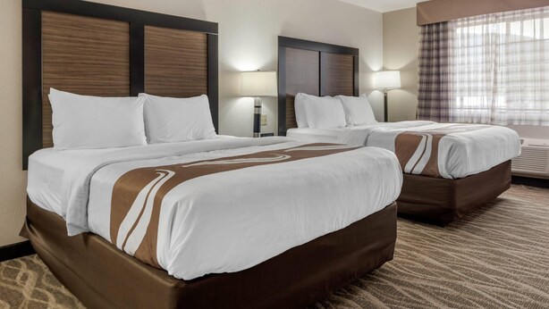 Habitación de hotel con dos camas, cada una con cabecero de madera, ropa de cama blanca y un detalle en marrón. Se ve una ventana con cortinas y una lámpara de pared.