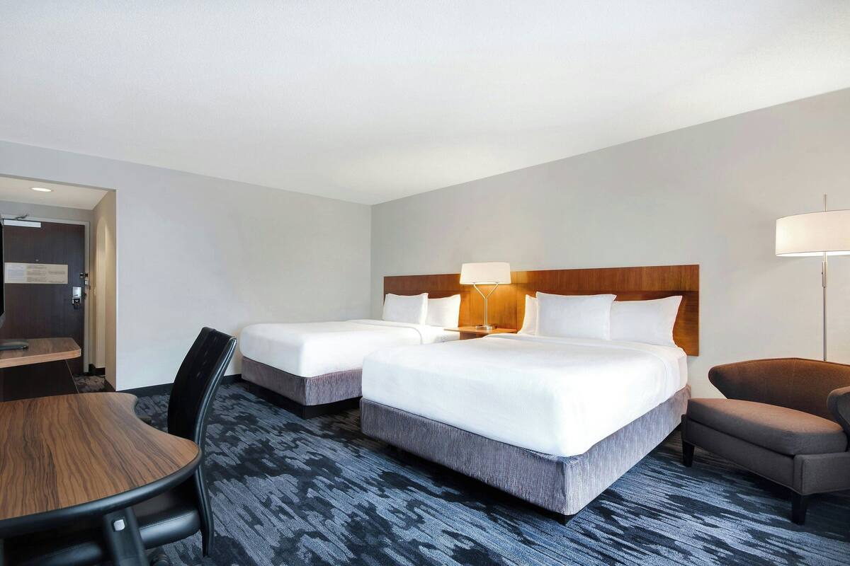 A hotel room with two beds, a desk, a chair, and a lamp.
