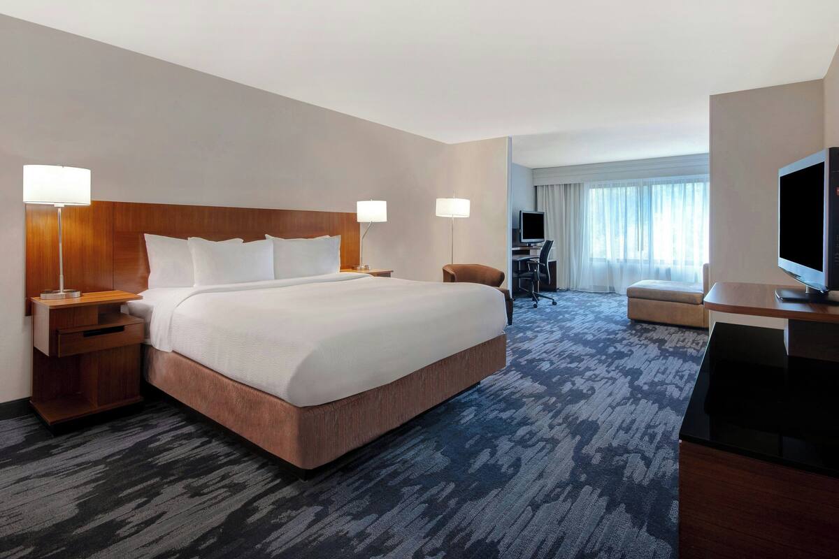 A hotel room with a large bed, two bedside tables with lamps, a desk with a TV, a chair, and a view of the city through large windows.