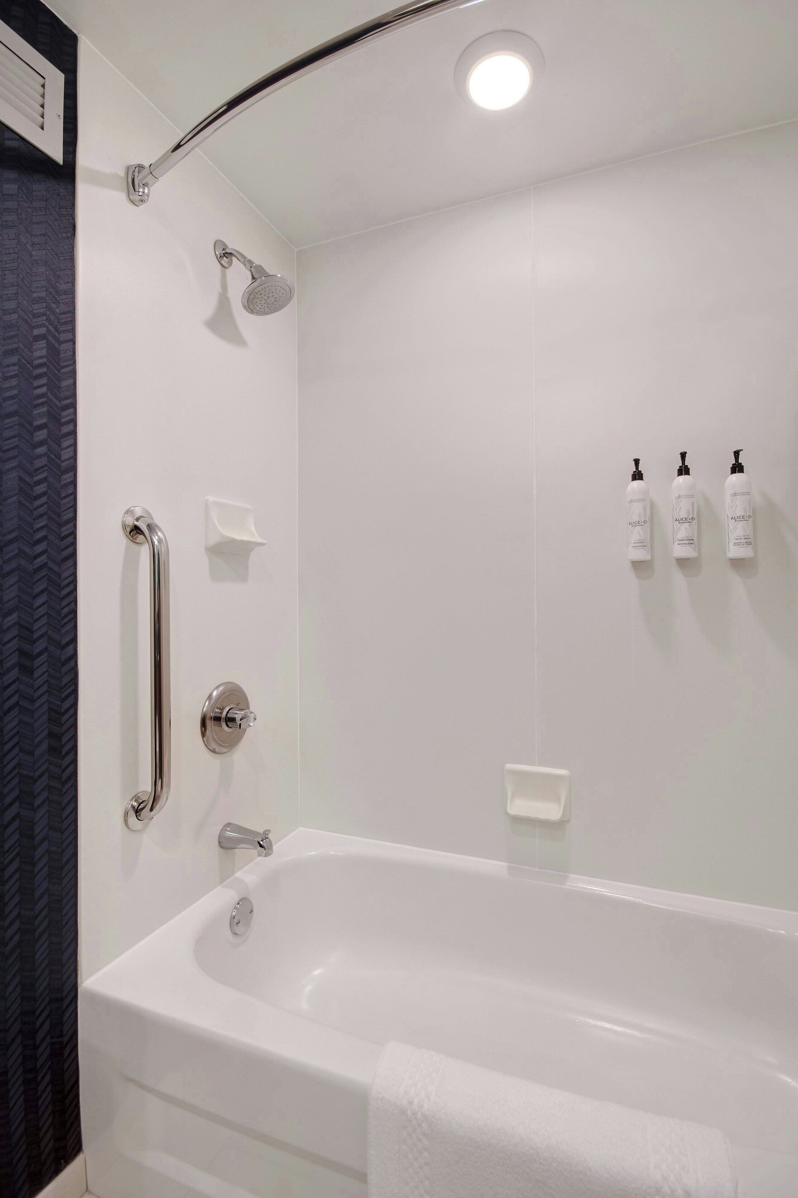 A modern bathroom with a white bathtub, a handheld showerhead, a handheld shower, and three bottles of shampoo on the wall.