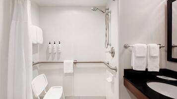 A modern bathroom with a shower, grab bars, a white chair, and a sink with a mirror.