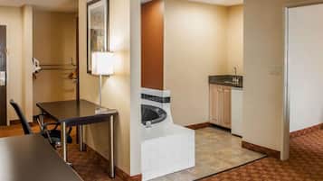 Quarto de hotel com mesa de jantar, pequeno espaço de cozinha, banheiro com banheira e espelho na parede.