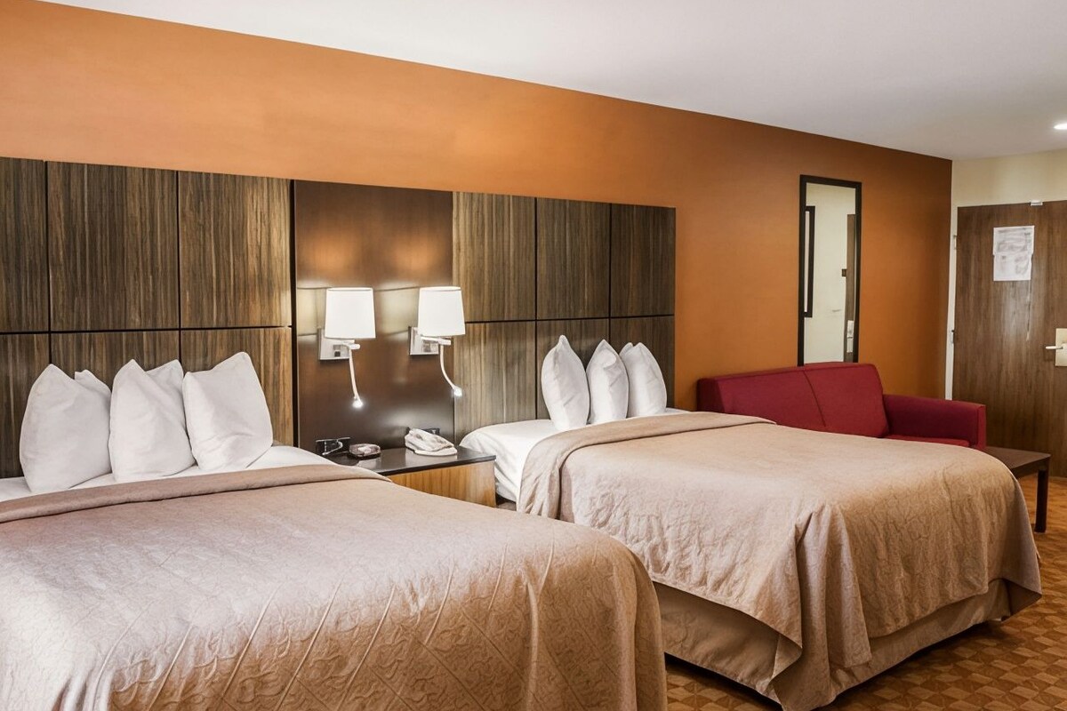 A hotel room with two beds, a red chair, a nightstand with a lamp, and a wooden headboard.