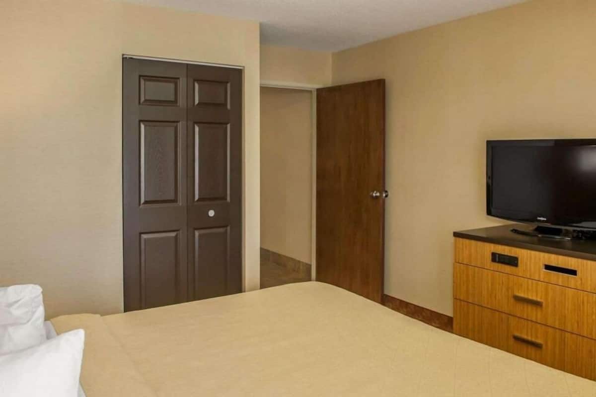 Quarto de hotel com cama, televisão, penteadeira de madeira e porta.