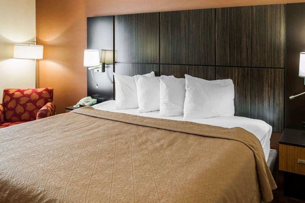 Quarto de hotel com uma cama grande, uma poltrona vermelha, mesa de cabeceira com abajur e telefone.