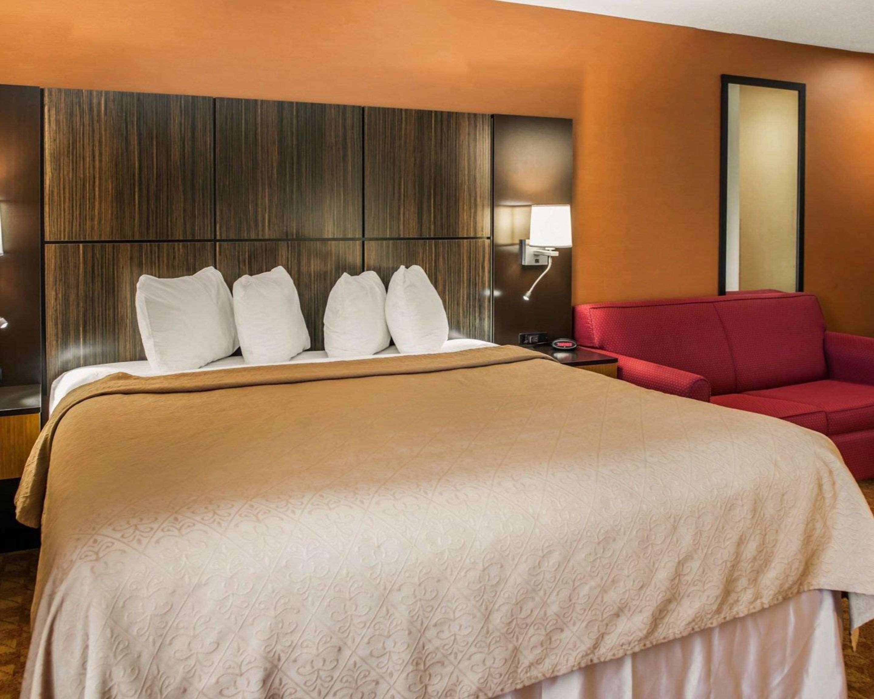 A hotel room with a large bed, a red sofa, a wooden headboard, and a wall-mounted lamp.