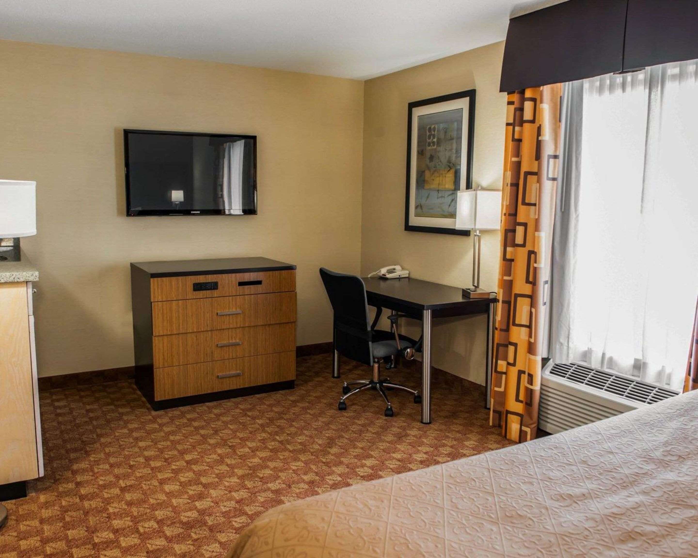 A hotel room with a bed, a desk with a chair, a dresser, a television, a framed picture, and a window with curtains.