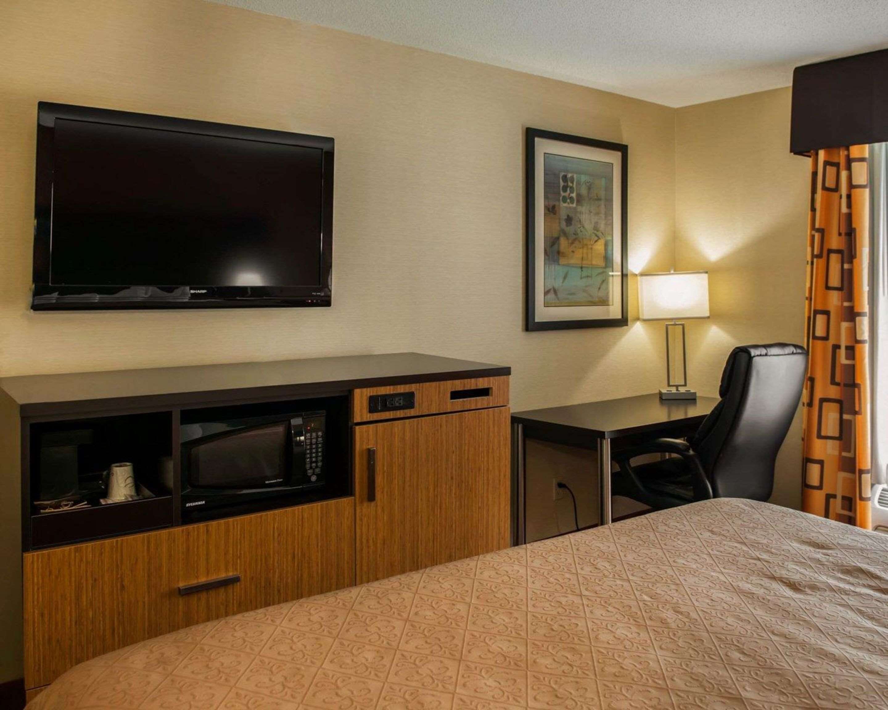 A hotel room with a bed, a desk with a computer, a television, a microwave, a chair, and a lamp.