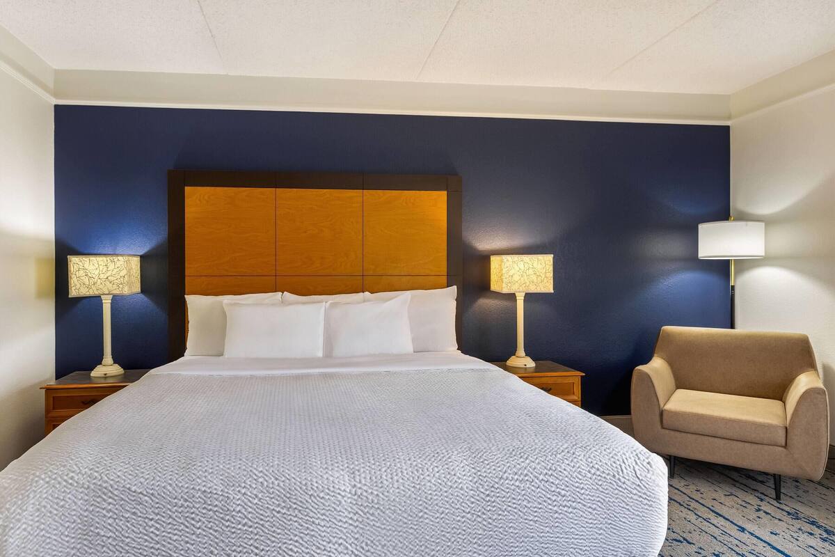 Ein Hotelzimmer mit einem großen Bett, zwei Nachttischen mit Lampen, einem Holzkopfteil und einem beigefarbenen Sessel.