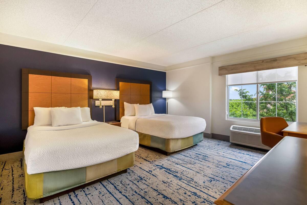 Ein Hotelzimmer mit zwei Betten, einem Schreibtisch, einem Sessel, einem Fenster mit Aussicht und einer Wand-Klimaanlage.