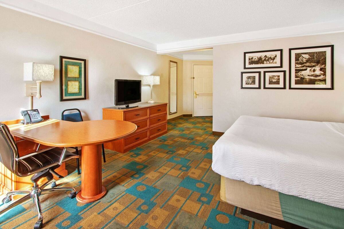 Ein Hotelzimmer mit einem Bett, einem Schreibtisch mit Stuhl, einem Fernseher, einem Schminktisch und gerahmten Bildern an der Wand.