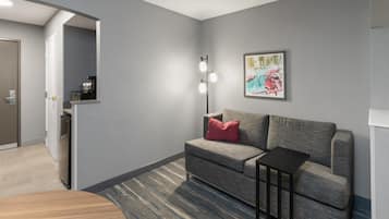 Una habitación de hotel moderna con un sofá gris, una mesita auxiliar pequeña, una alfombra estampada y un cuadro en la pared.