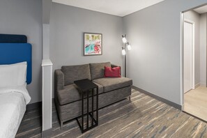 Una habitación de hotel con un sofá gris, una mesita de noche negra pequeña, un cuadro en la pared y una puerta blanca.