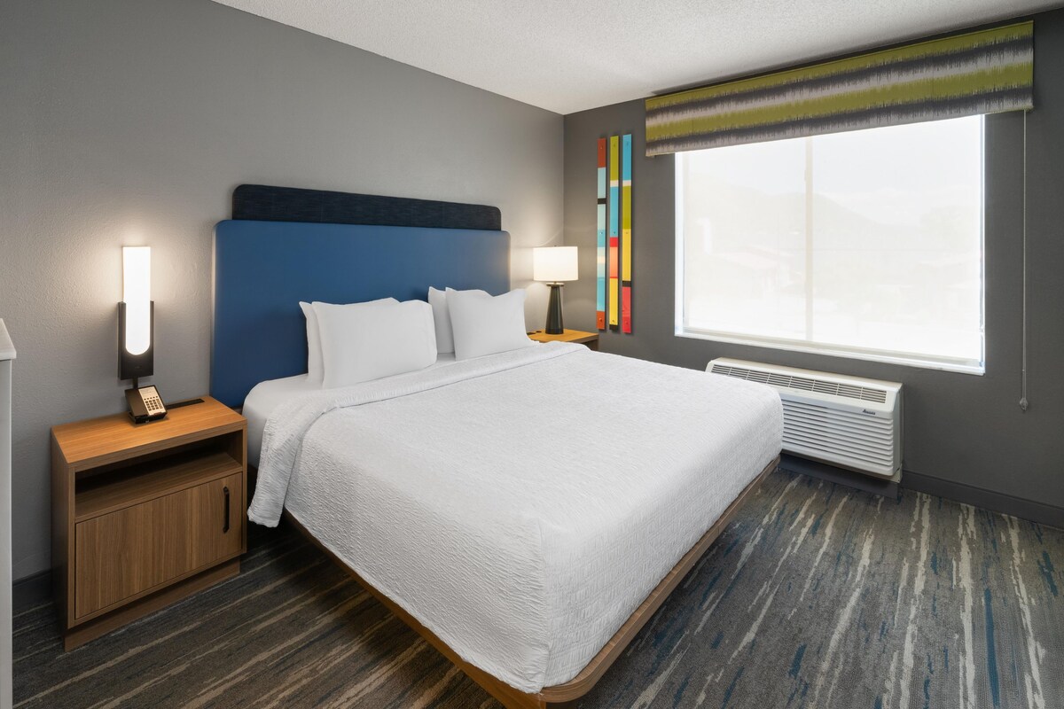 A hotel room with a bed, bedside tables, a lamp, a window with blinds, and a wall-mounted air conditioner.