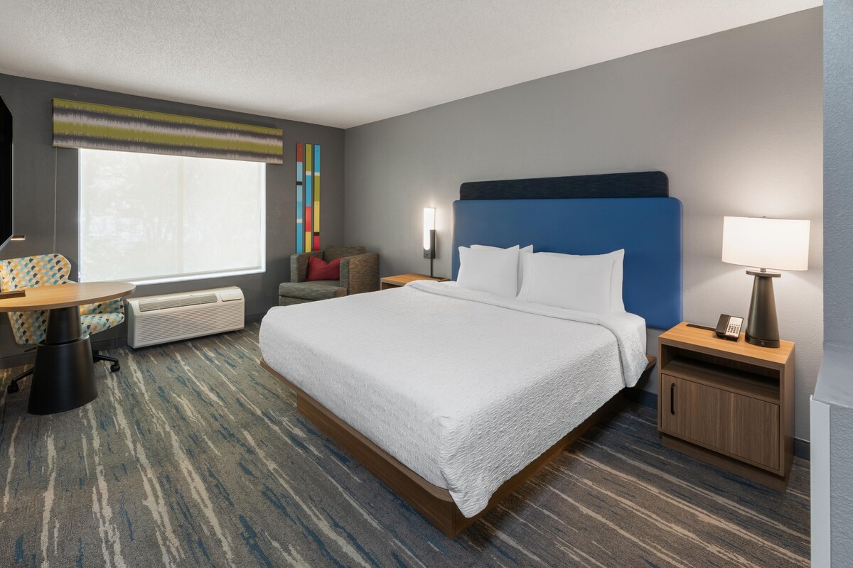A hotel room with a large bed, a desk with a chair, a window, a wall-mounted lamp, and a bedside table with a lamp.
