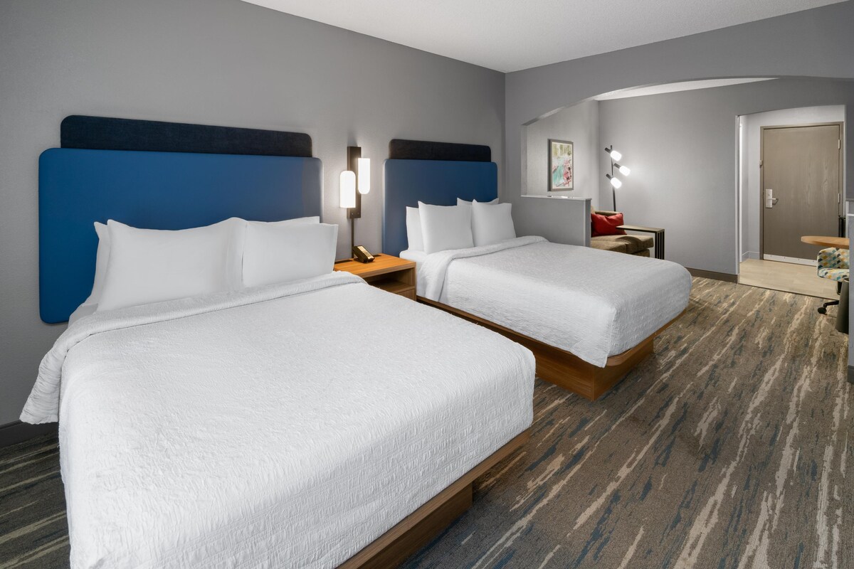 A hotel room with two beds, a nightstand with a lamp, a small table, and a door leading to another room.