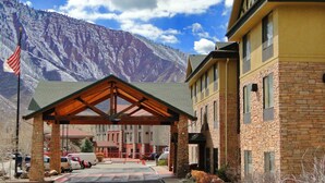 Entrada de un hotel con zona cubierta, montañas al fondo y aparcamiento.
