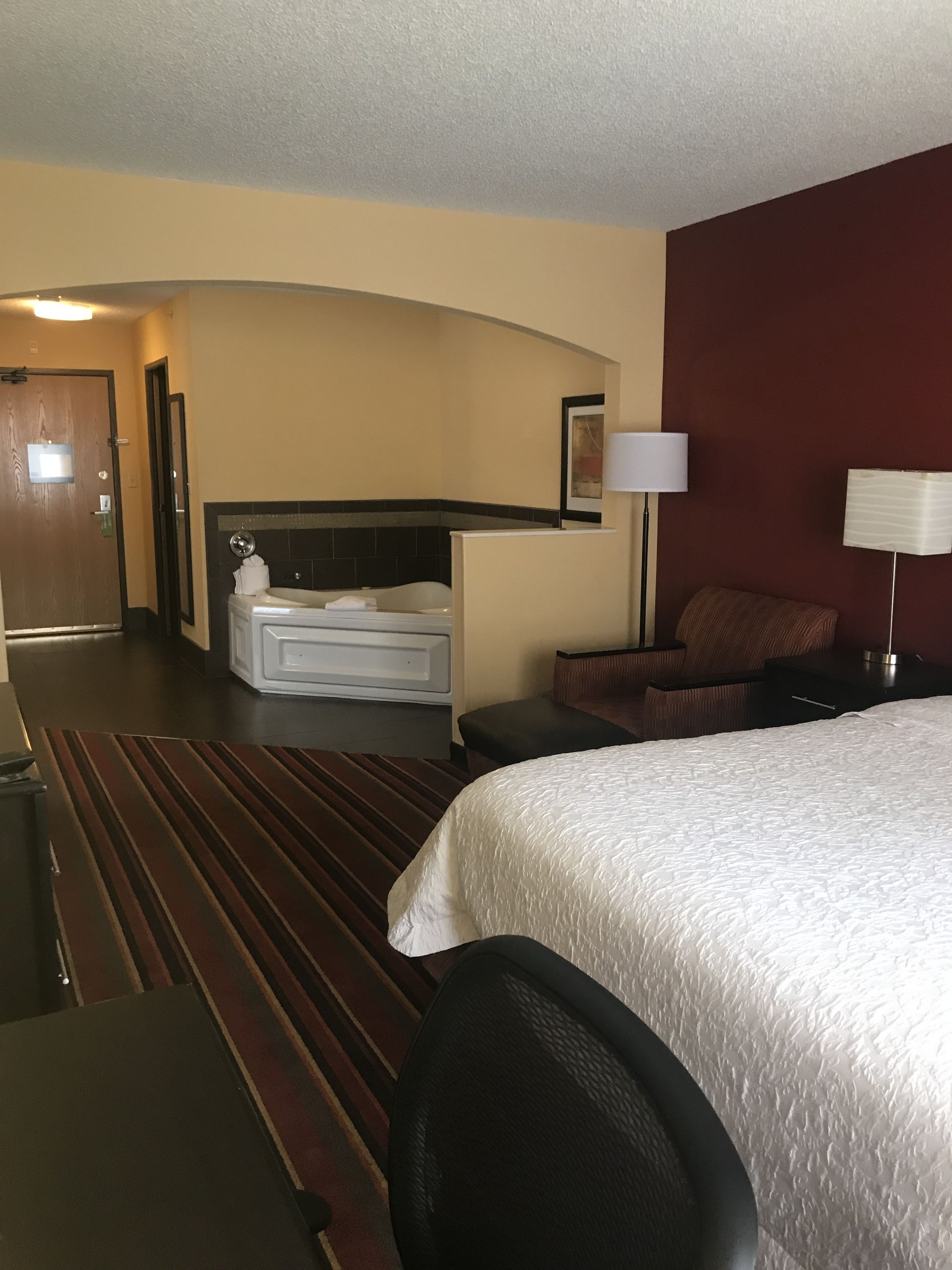 A hotel room with a bed, a desk, a chair, a sofa, a nightstand, a lamp, a bathroom with a bathtub, and a door.