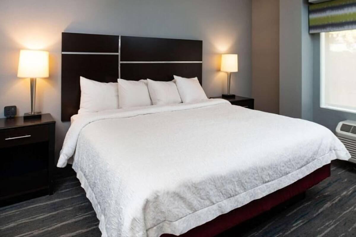 Habitación de hotel con una cama grande, dos mesitas de noche con lámparas, un cabecero oscuro y una ventana con persianas.