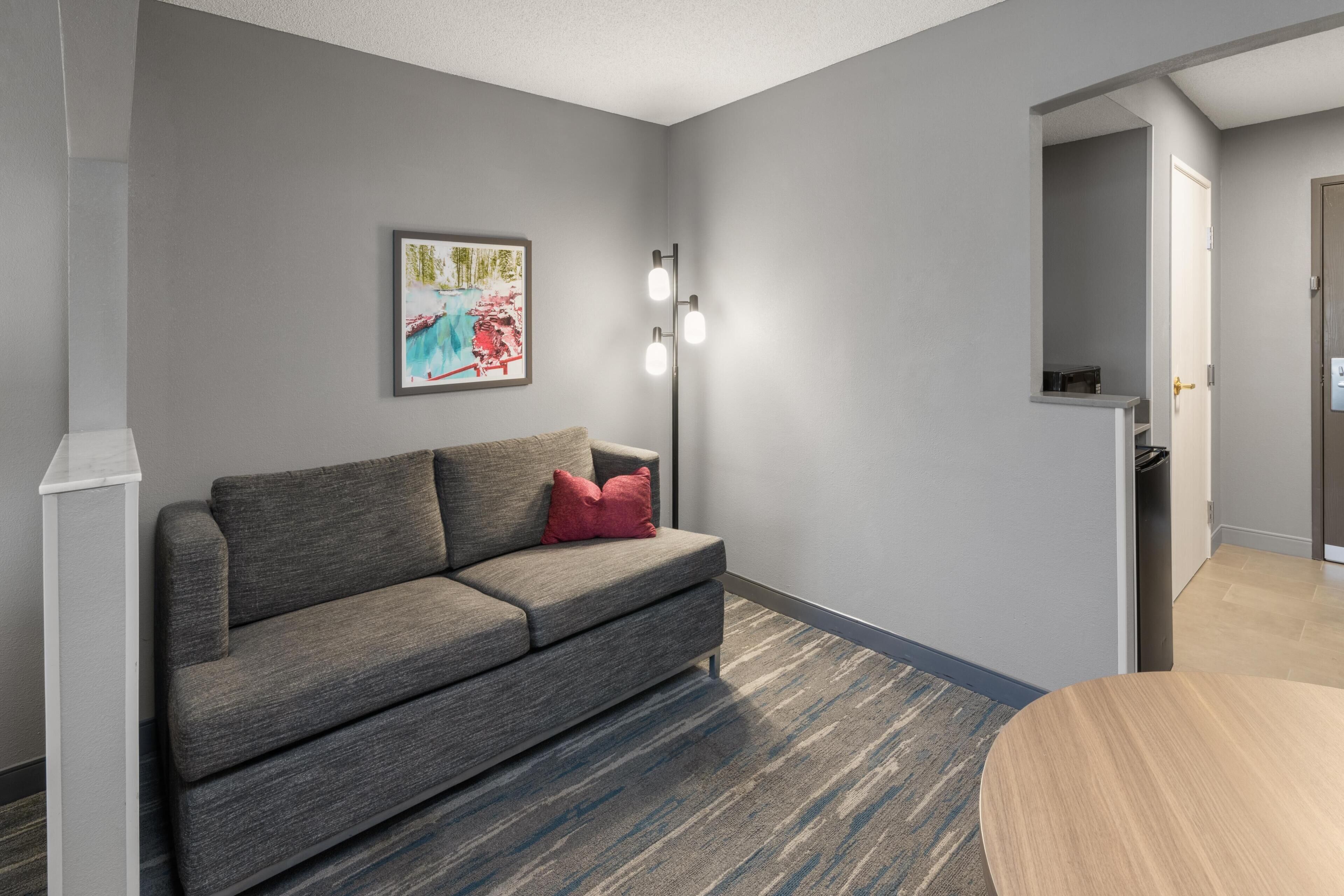 Ein modernes Wohnzimmer mit einem grauen Sofa, einem roten Kissen, einem gerahmten Bild an der Wand und einer Stehlampe.