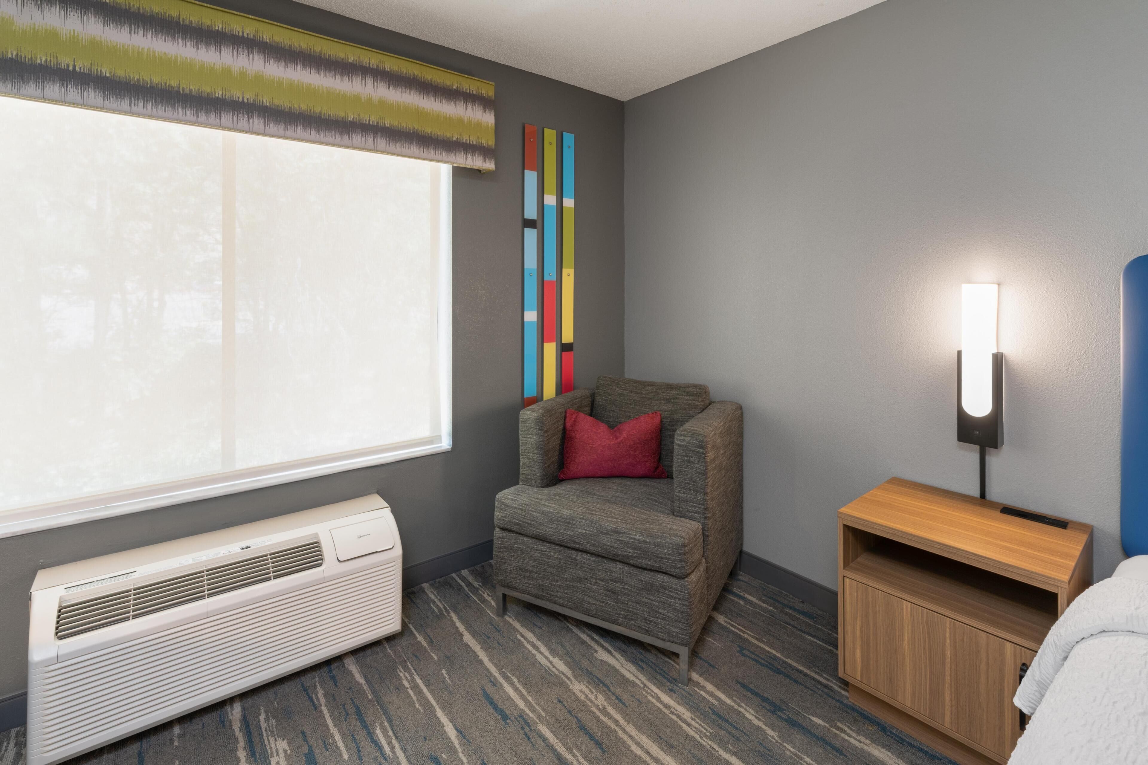 A hotel room with a gray sofa, a standing heater, a bedside table with a lamp, and a window with a view.