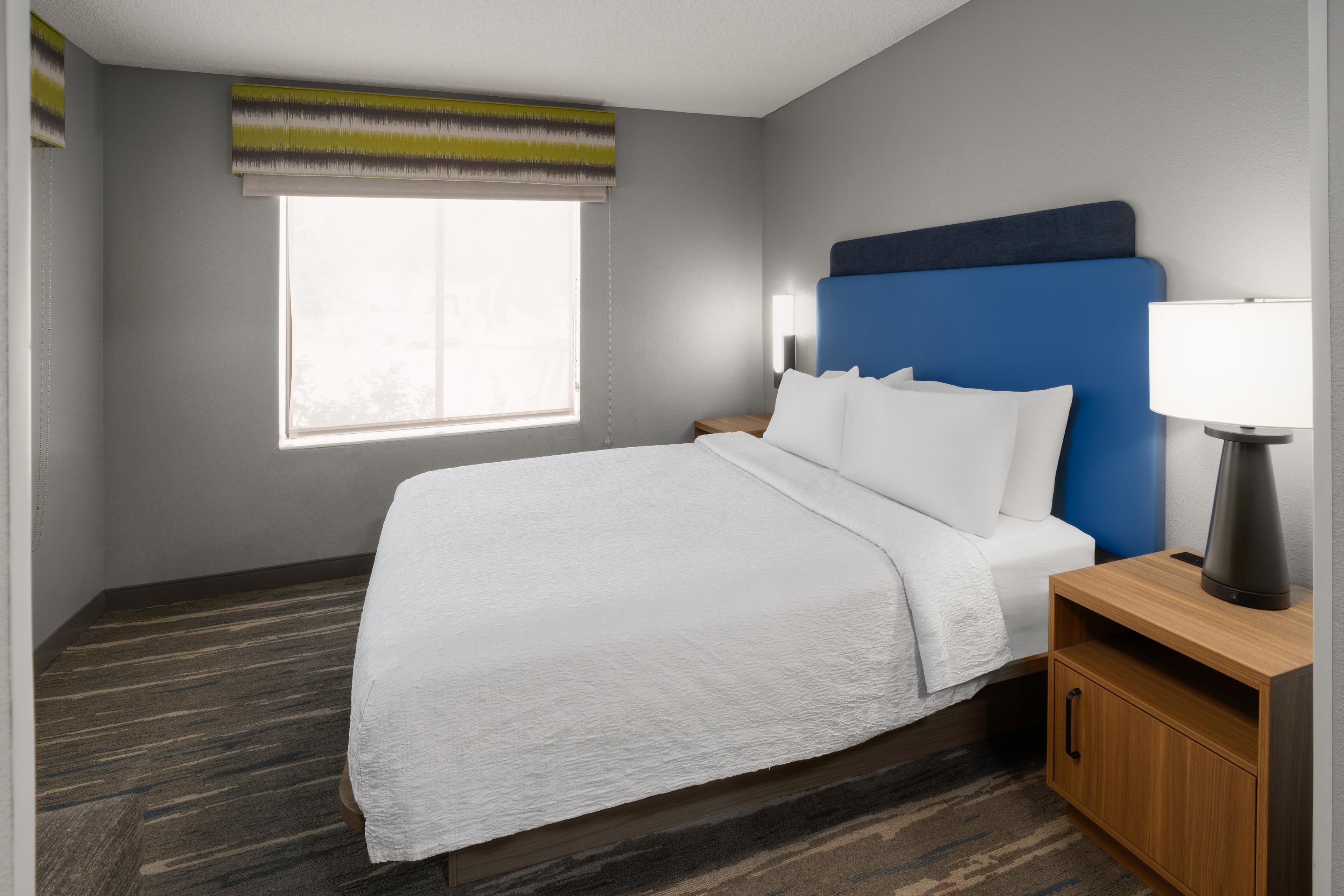A hotel room with a large bed, a nightstand with a lamp, a window with blinds, and a carpeted floor.
