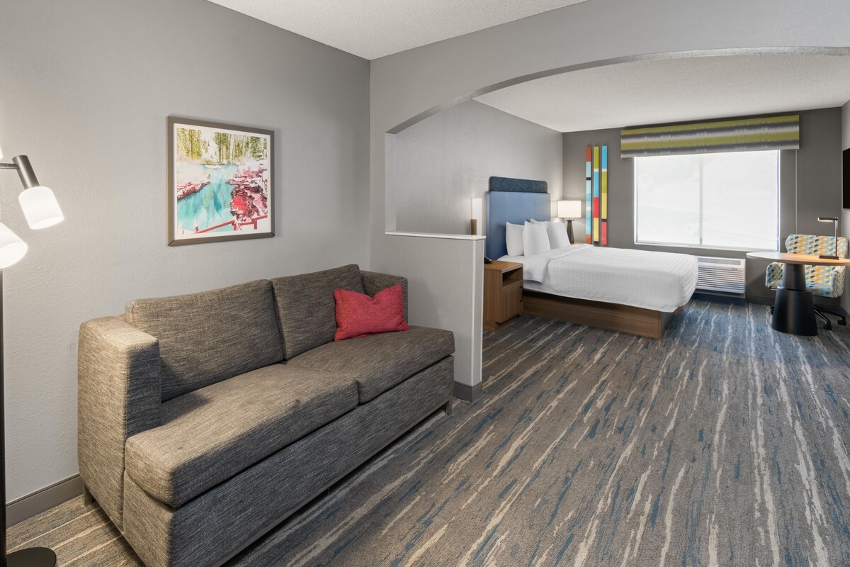 A hotel room with a sofa, a bed, a nightstand, a lamp, a window with a view, and a painting on the wall.