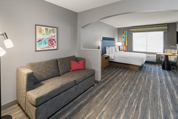A hotel room with a sofa, a bed, a nightstand, a lamp, a window with a view, and a painting on the wall.
