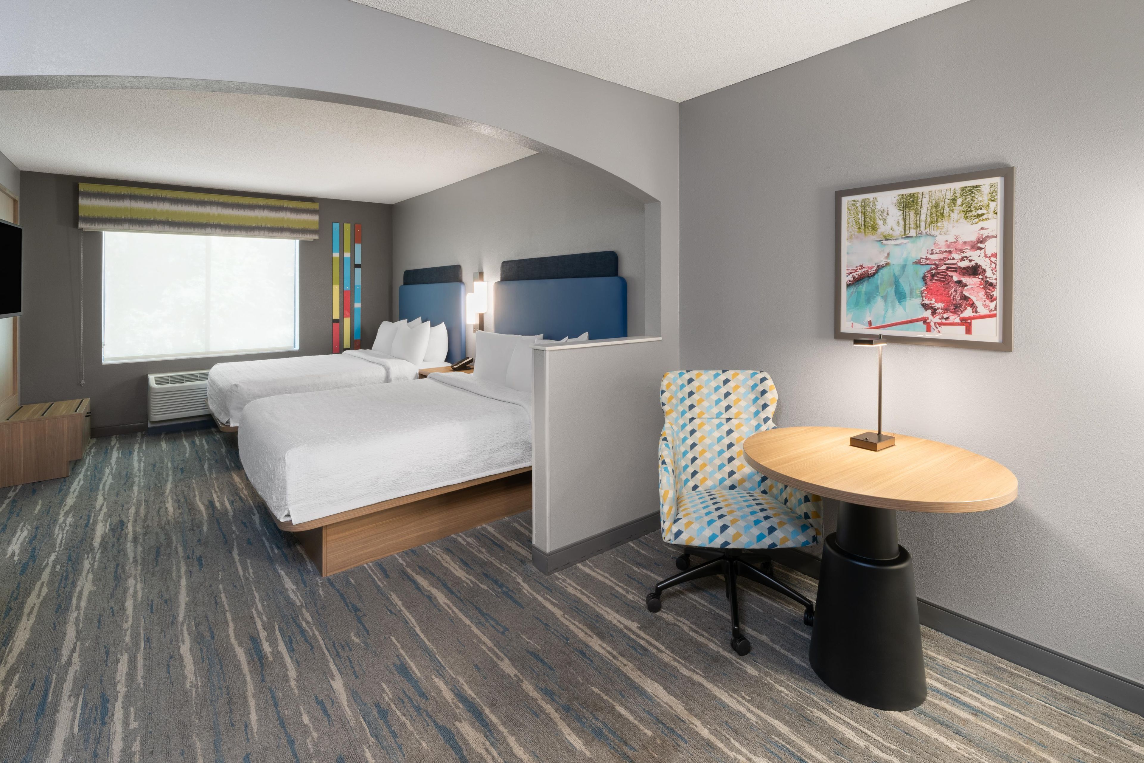 A hotel room with a bed, a desk, a chair, a window, and a painting on the wall.