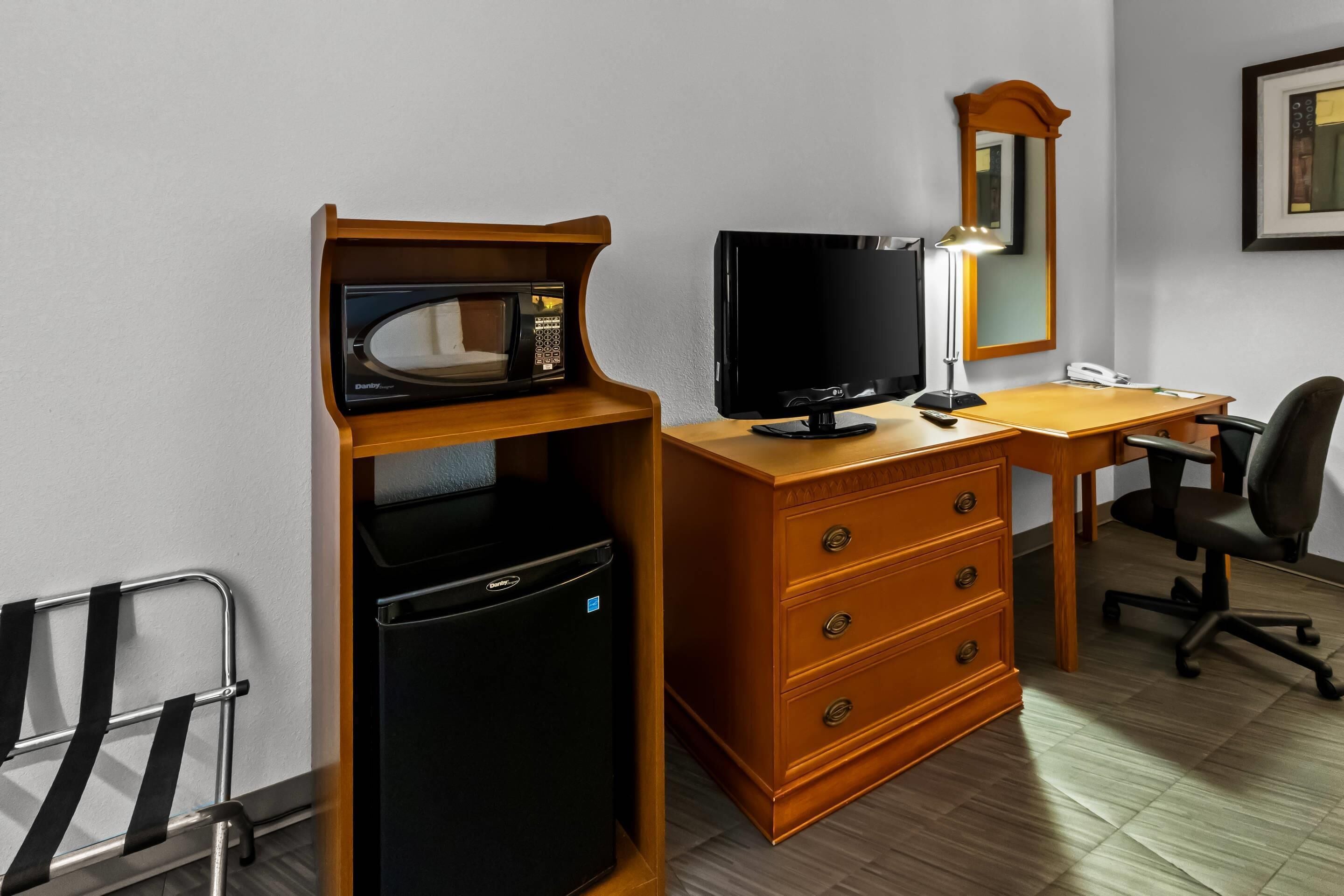 Une chambre d’hôtel compacte comprenant un bureau en bois, un réfrigérateur noir, une télévision, une lampe de bureau, une chaise et une petite étagère en métal.