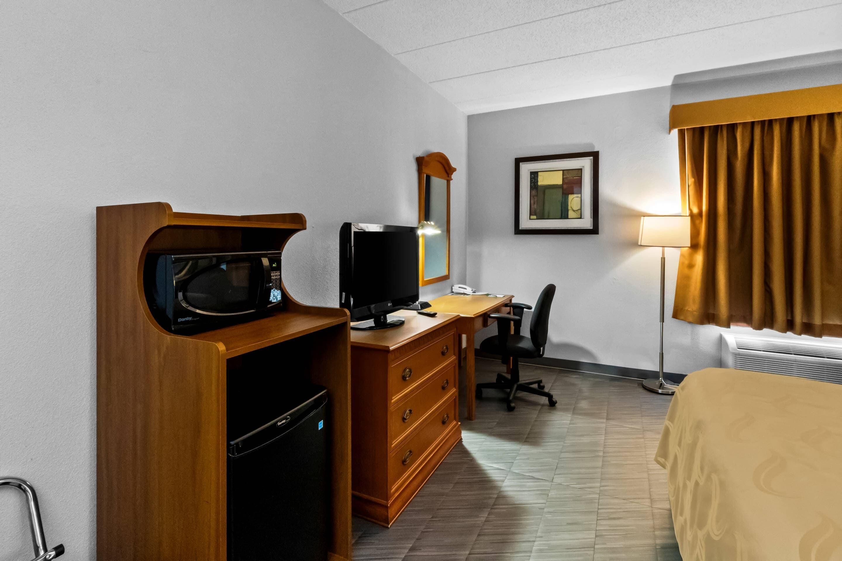 Habitación de hotel con escritorio de madera, microondas, televisión, silla, cama, lámpara y un cuadro en la pared.