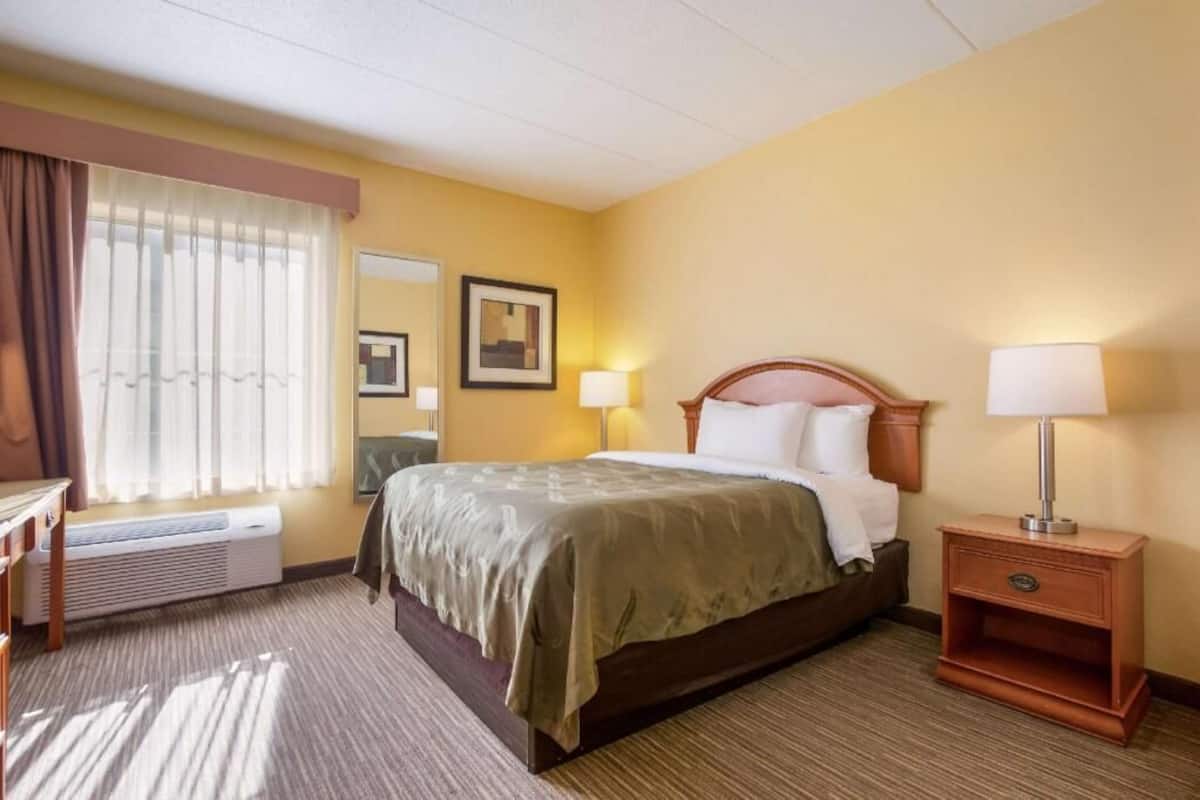 A hotel room with a bed, bedside lamps, a window with curtains, a radiator, and a wall-mounted picture.